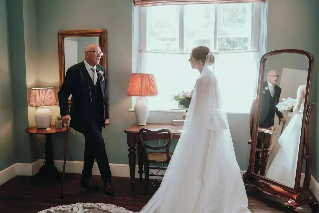 Bride in her wedding dress and veil standing by a window speaking with her father, who holds a walking cane, during a quiet pre-ceremony moment indoors.
