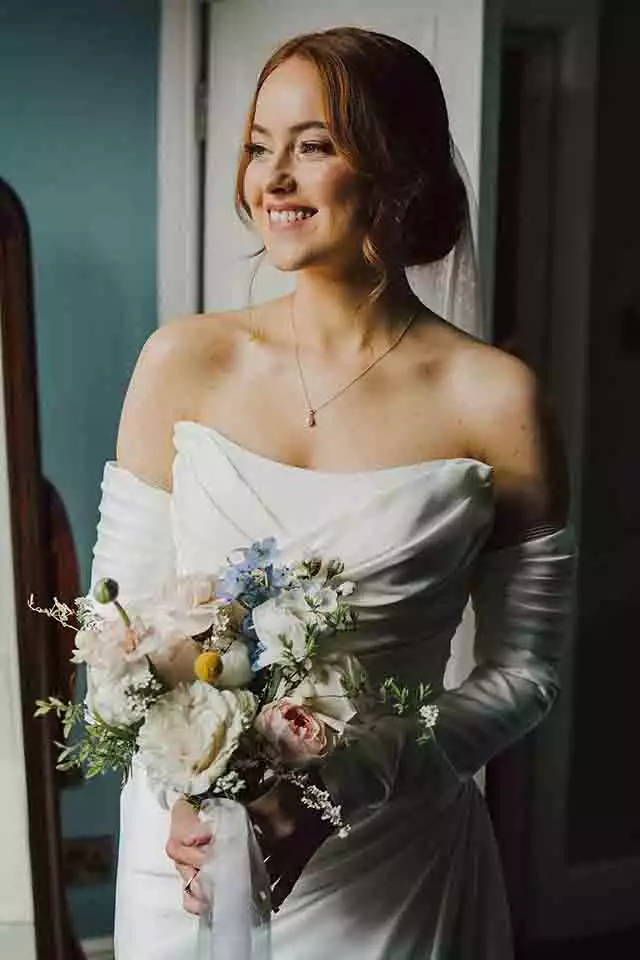 Bride in an elegant off-the-shoulder wedding dress holding a bouquet of pastel flowers while smiling softly near a window during wedding preparations.