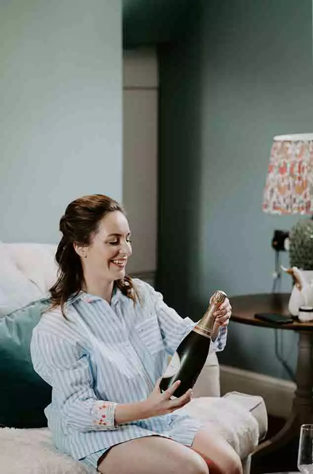 Bride in striped pajamas sitting on a sofa and smiling while opening a bottle of champagne during relaxed wedding morning preparations.