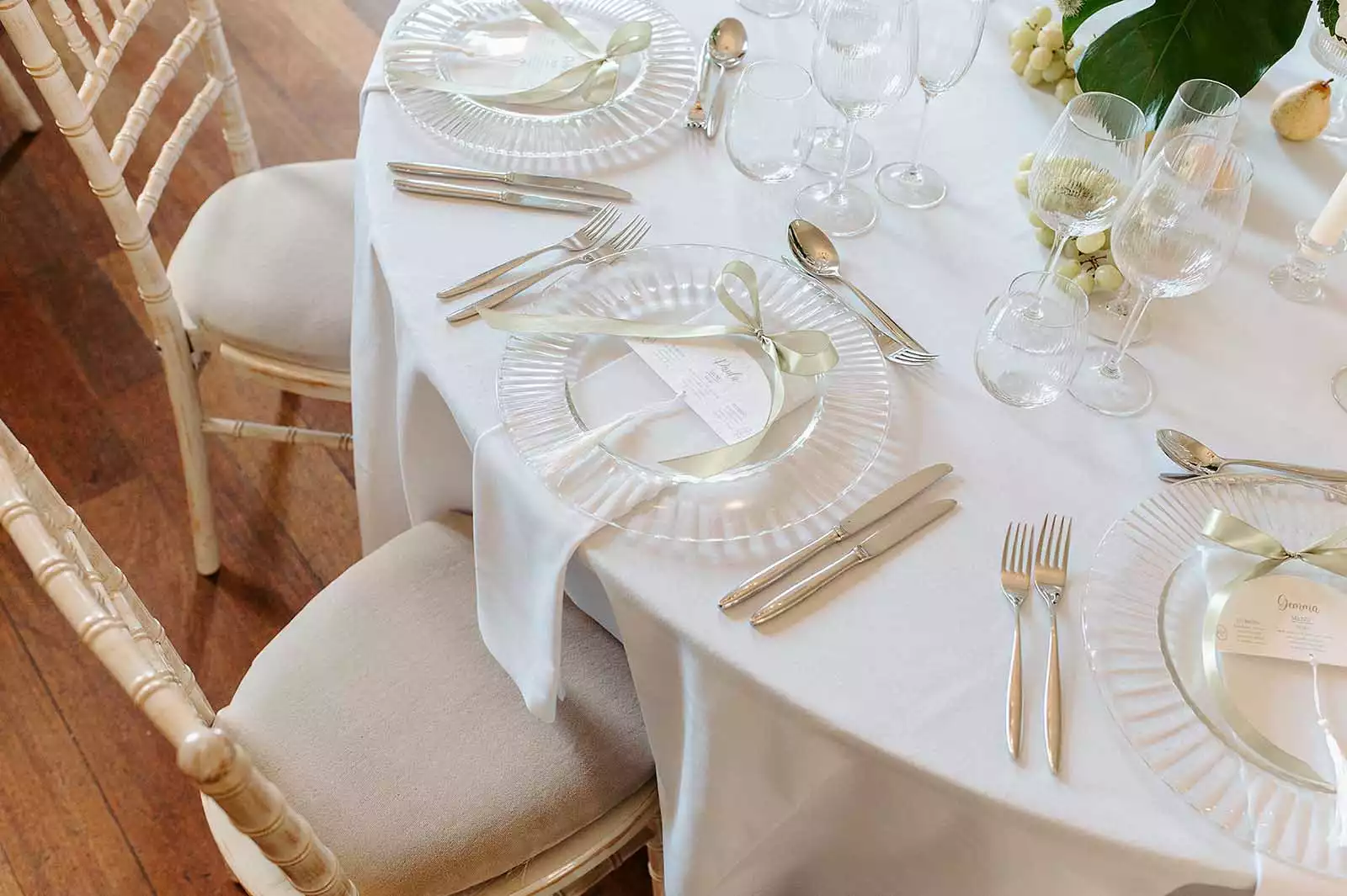 Elegant wedding reception table set with transparent glass plates, gold cutlery, folded ribbon napkins, and multiple wine glasses on a white tablecloth, with chiavari chairs and decorative greenery.