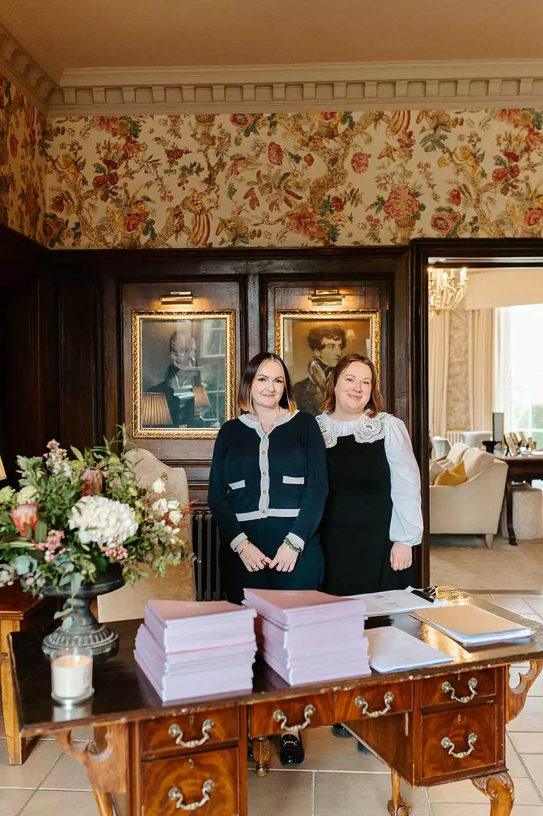 Two wedding venue staff members standing behind a desk with guest books and flowers in an elegant reception area.
