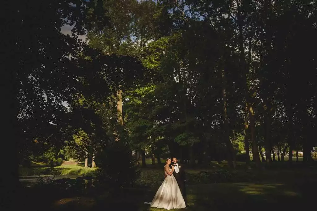 Bride and groom standing together beneath tall trees in the gardens at Rowton Castle, with warm evening light filtering through the woodland.