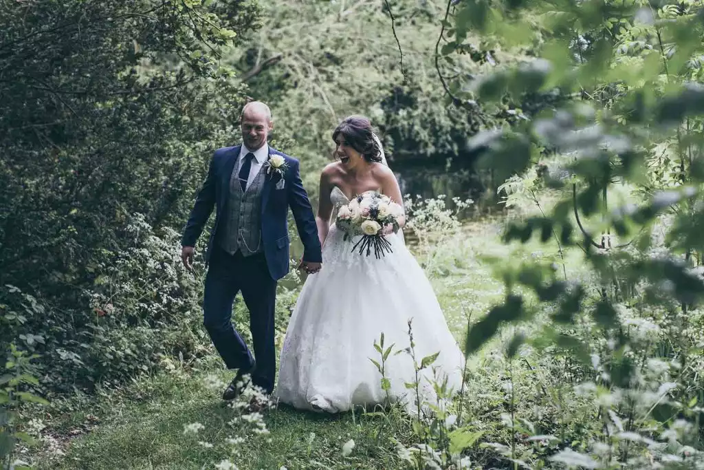 Bride holding a bouquet and groom in a blue suit walking hand in hand along a leafy lakeside path in the natural woodland grounds of Rowton Castle.