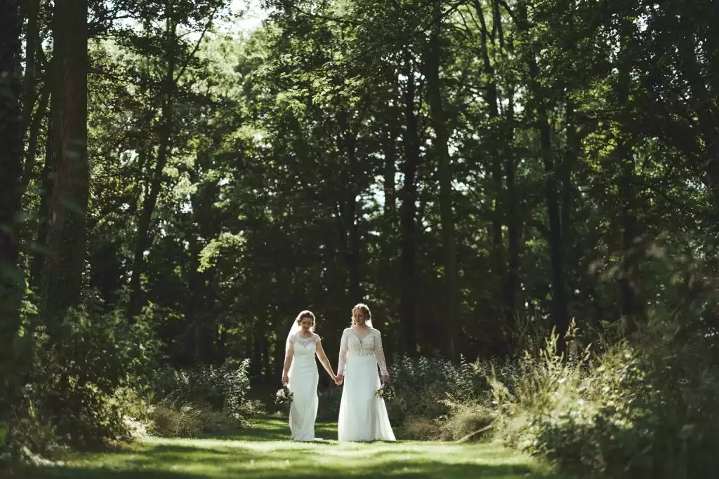 Two brides in wedding dresses walking hand in hand along a sunlit woodland path at Rowton Castle, surrounded by tall trees and soft green foliage.